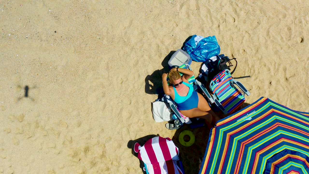 Woman in blue swimsuit adjusts near her head while lying beside a striped umbrella.