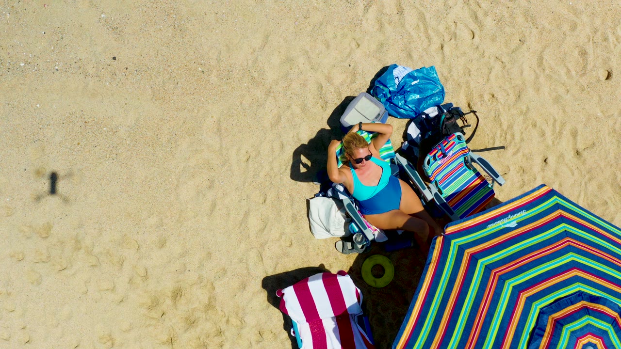 At two seconds, the La Laguna Beach Los Cabos overhead frame holds on the woman in the blue swimsuit, with the colorful umbrella edge lower right and beach bags above her on pale sand.