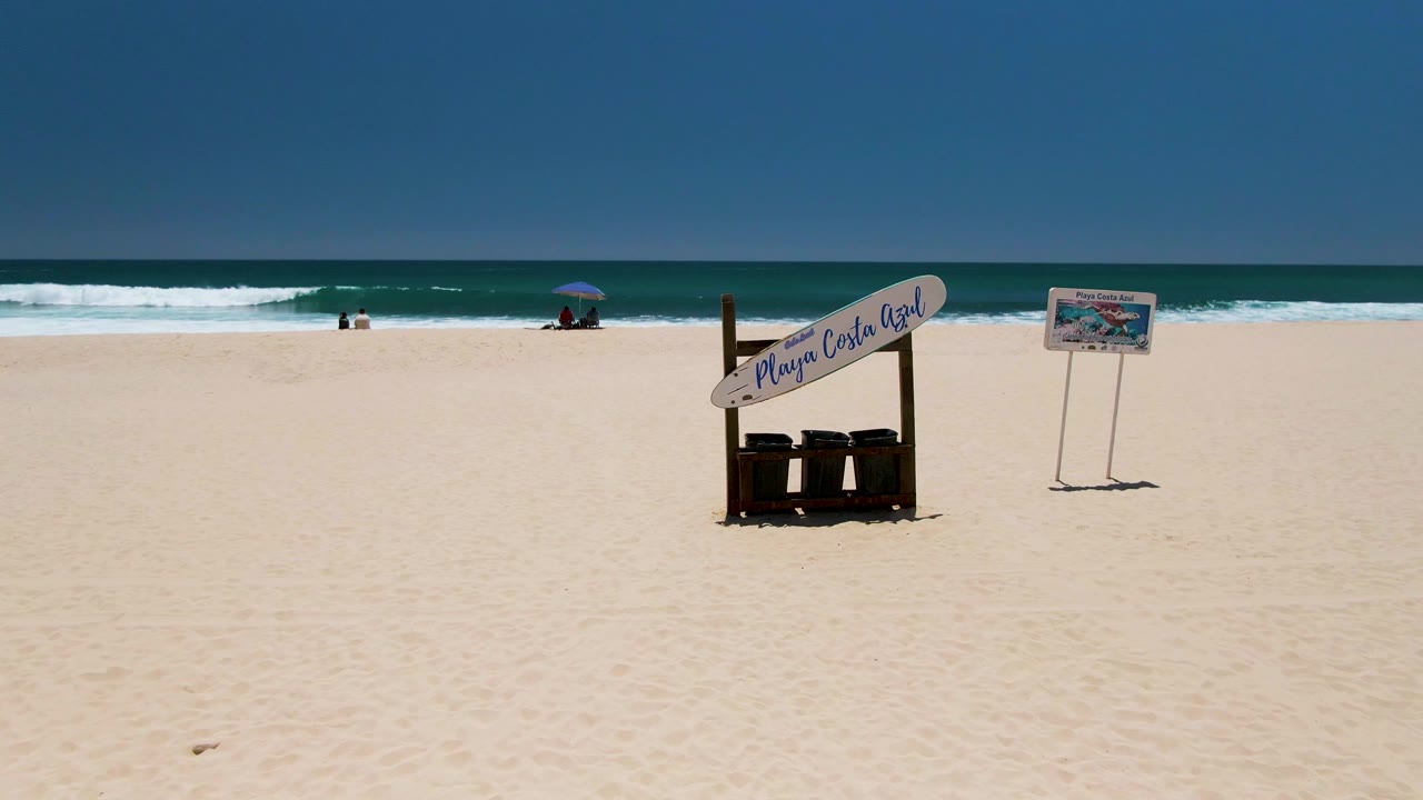 Costa Azul Beach in Los Cabos with broad pale sand, a Playa Costa Azul sign structure and nearby signboard, distant surf, small beachgoers, and a blue umbrella near the shoreline.