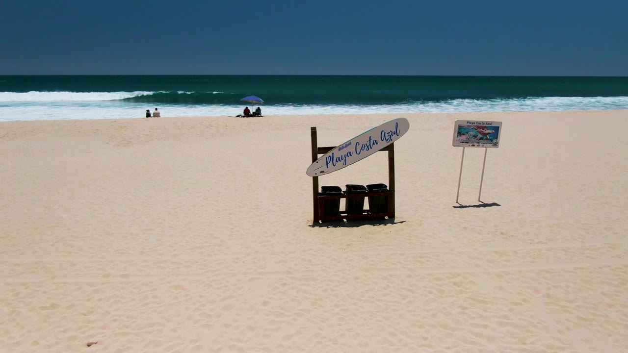 Sandy beach with Playa Costa Azul sign, separate signboard, waves, and small figures near the water.