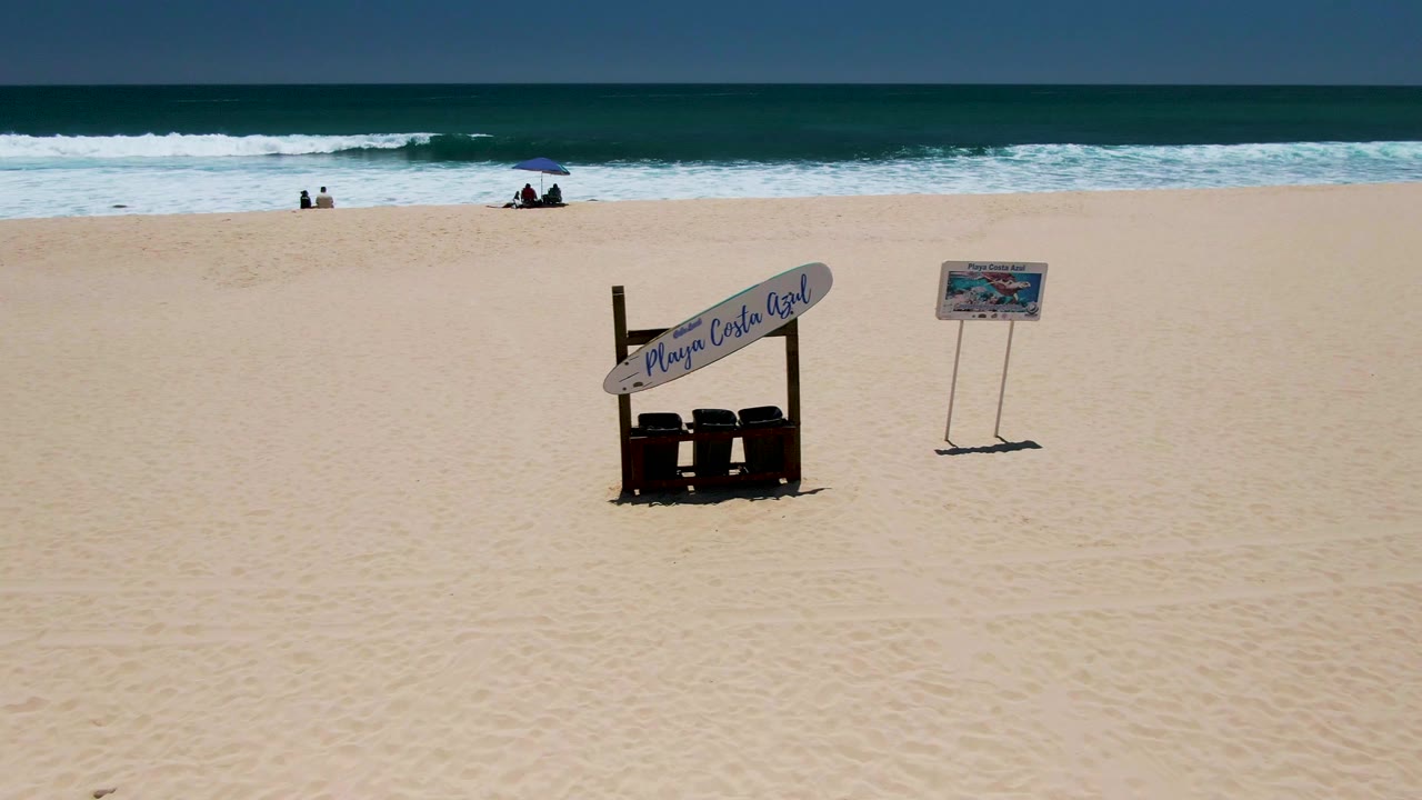Costa Azul Beach in Los Cabos at 0:02, with the Playa Costa Azul sign structure and freestanding signboard on the sand while beachgoers remain small near the breaking waves.