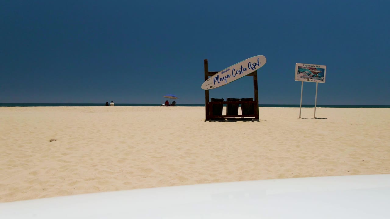Wide view of Costa Azul Beach in Los Cabos with pale sand, the Playa Costa Azul sign near center, a smaller beach sign, distant people near the waterline, and a white foreground edge at the bottom.