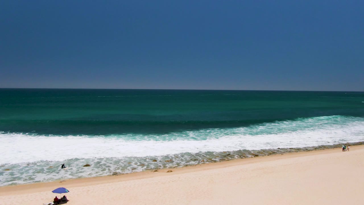 Aerial view of Costa Azul Beach in Los Cabos, Mexico, with deep blue ocean, white surf, pale sand, a blue umbrella, and small beachgoers near the lower edge.