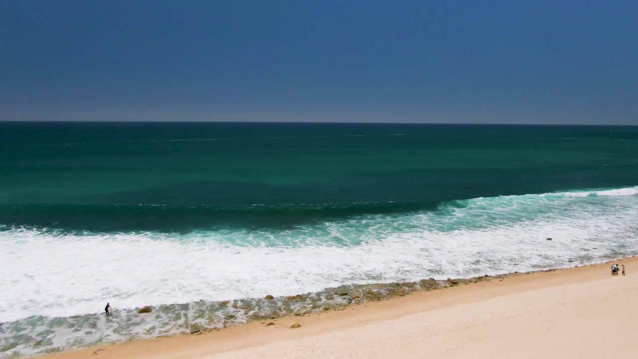 Aerial view of Costa Azul Beach in Los Cabos, Mexico, with dark blue water beyond the break, foamy surf across the middle, sand below, and two small beachgoers near the right edge.