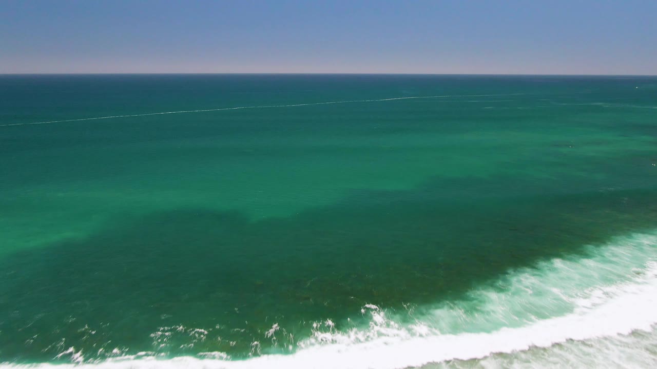 Aerial view at Costa Azul Beach with green ocean filling the frame, a curved white wave line near the bottom, and a faint horizon under clear sky with no visible people.