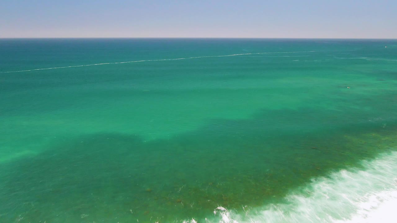 Aerial frame over Costa Azul Beach showing brighter turquoise water, faint seabed color, and white surf pushing in from the lower right with no visible people.
