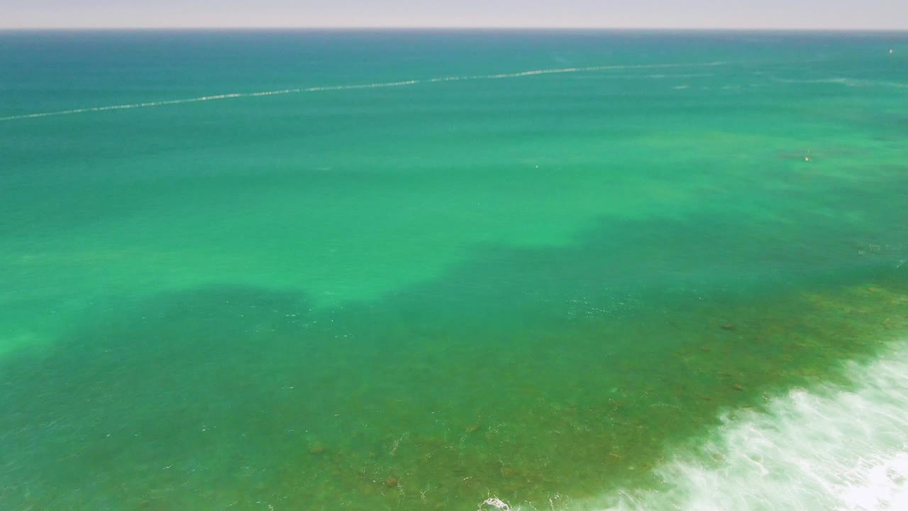 Aerial view at Costa Azul Beach with clear turquoise water, green seabed patches, a thin white foam line along the lower right, and no visible people.