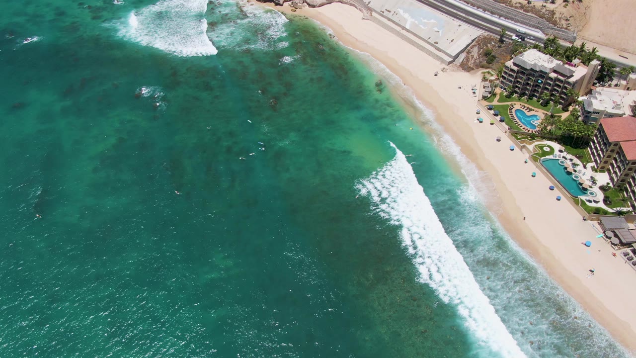 Opening aerial frame over Costa Azul Beach, Los Cabos, with turquoise water, a white breaking wave, pale sand, beachfront pools, and small distant beachgoers near the buildings.