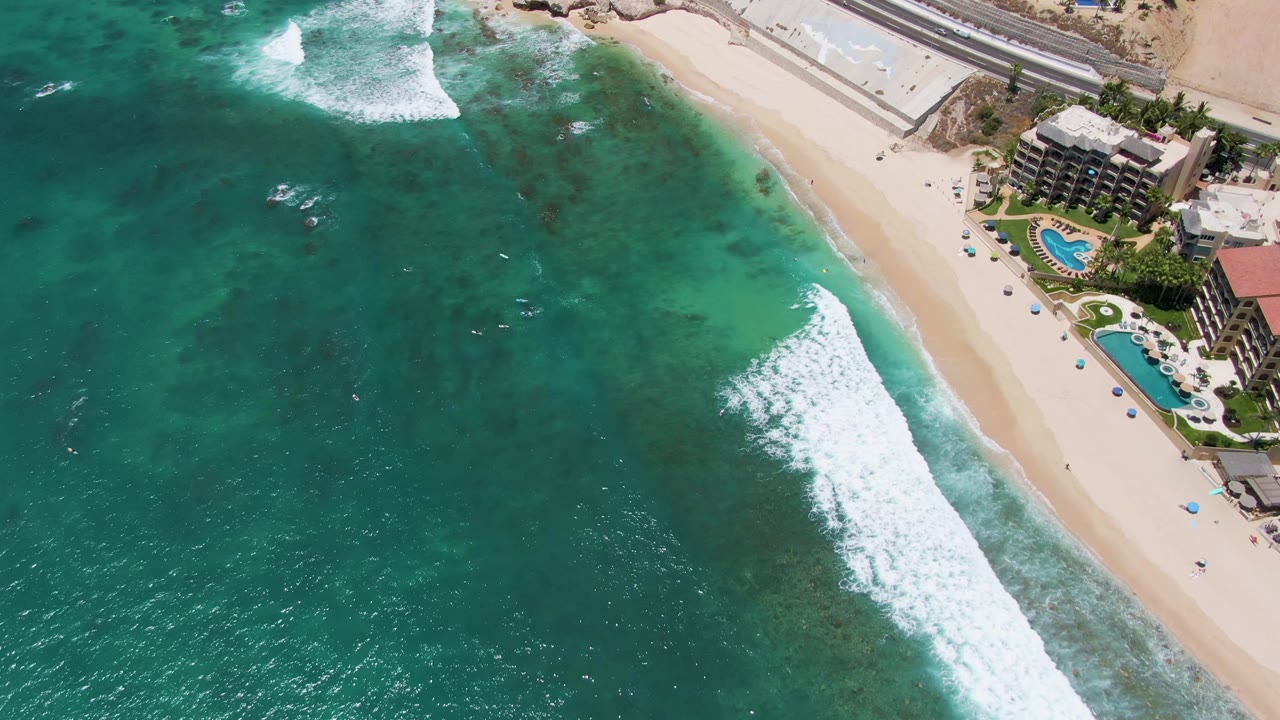 Costa Azul Beach aerial frame with a compact white wave breaking near the shoreline, darker patches in the clear water, pale sand, and pool areas beside coastal buildings.