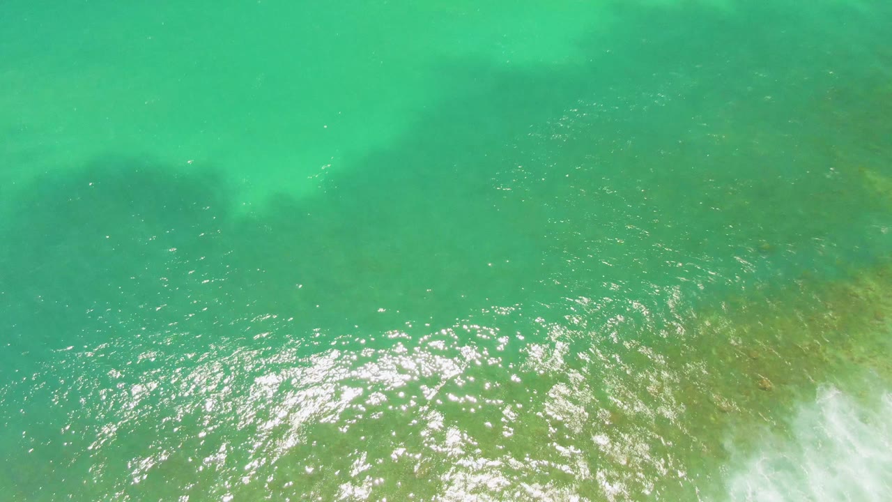 Aerial frame over Costa Azul Beach water in Los Cabos, showing a broader patch of sun glitter across rippled turquoise surface, greenish seabed tones below, and no visible people.
