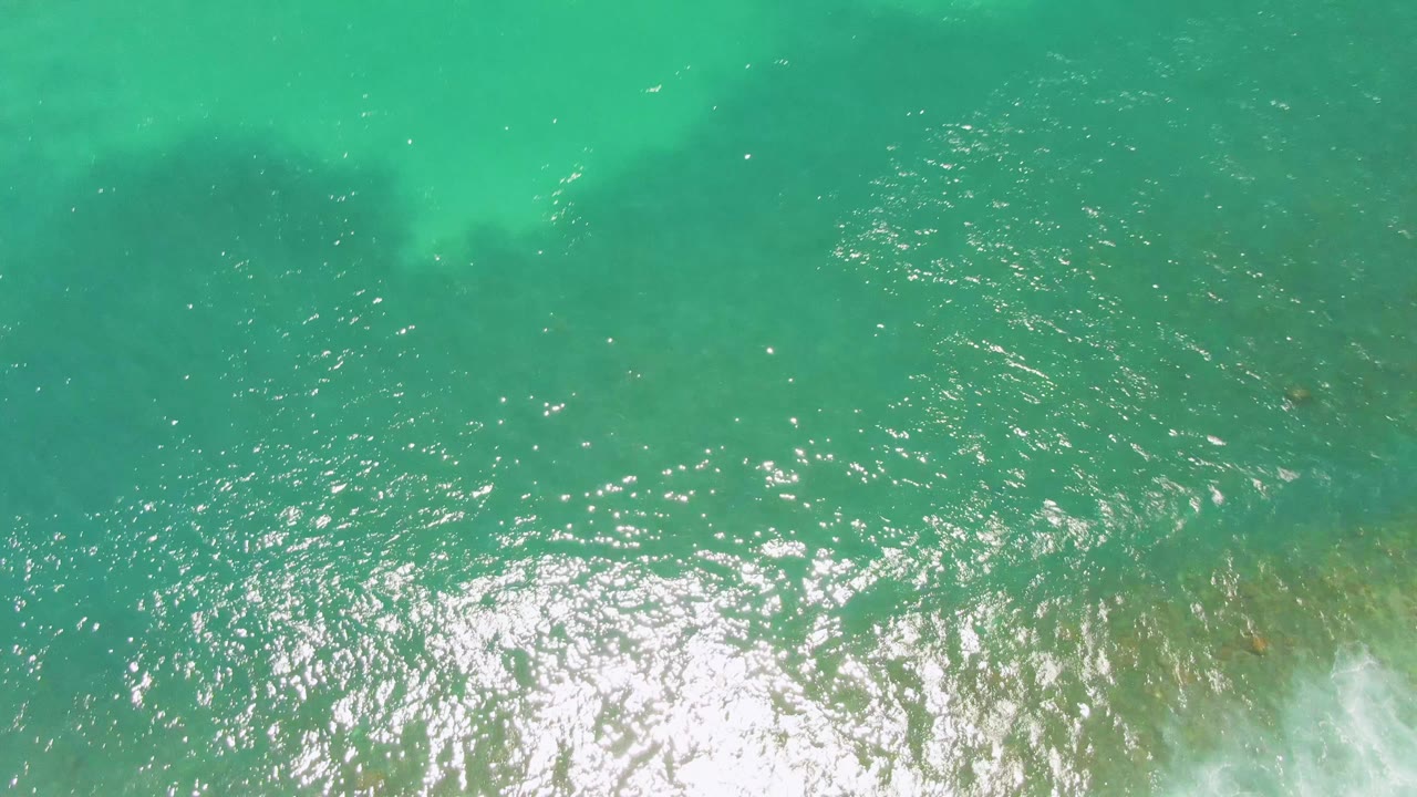 Top-down Costa Azul Beach ocean view with a dense round cluster of reflected sunlight on blue-green ripples, faint seabed patches at the edges, and no visible people.