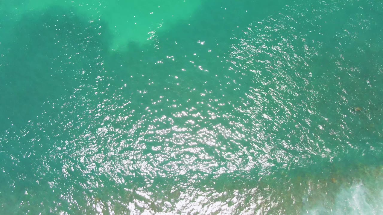 Costa Azul Beach water seen from above, with darker turquoise surface, scattered sunlight points, faint underwater color variation, a small strip of white surf near the bottom, and no visible people.
