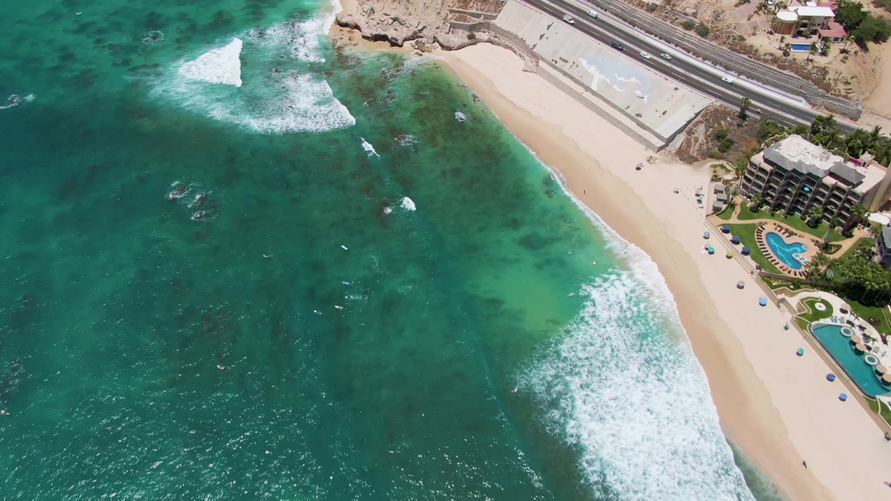 Aerial frame over Costa Azul Beach in Los Cabos showing turquoise water, white surf on pale sand, a coastal road, adjacent buildings with pools, and no visible people.
