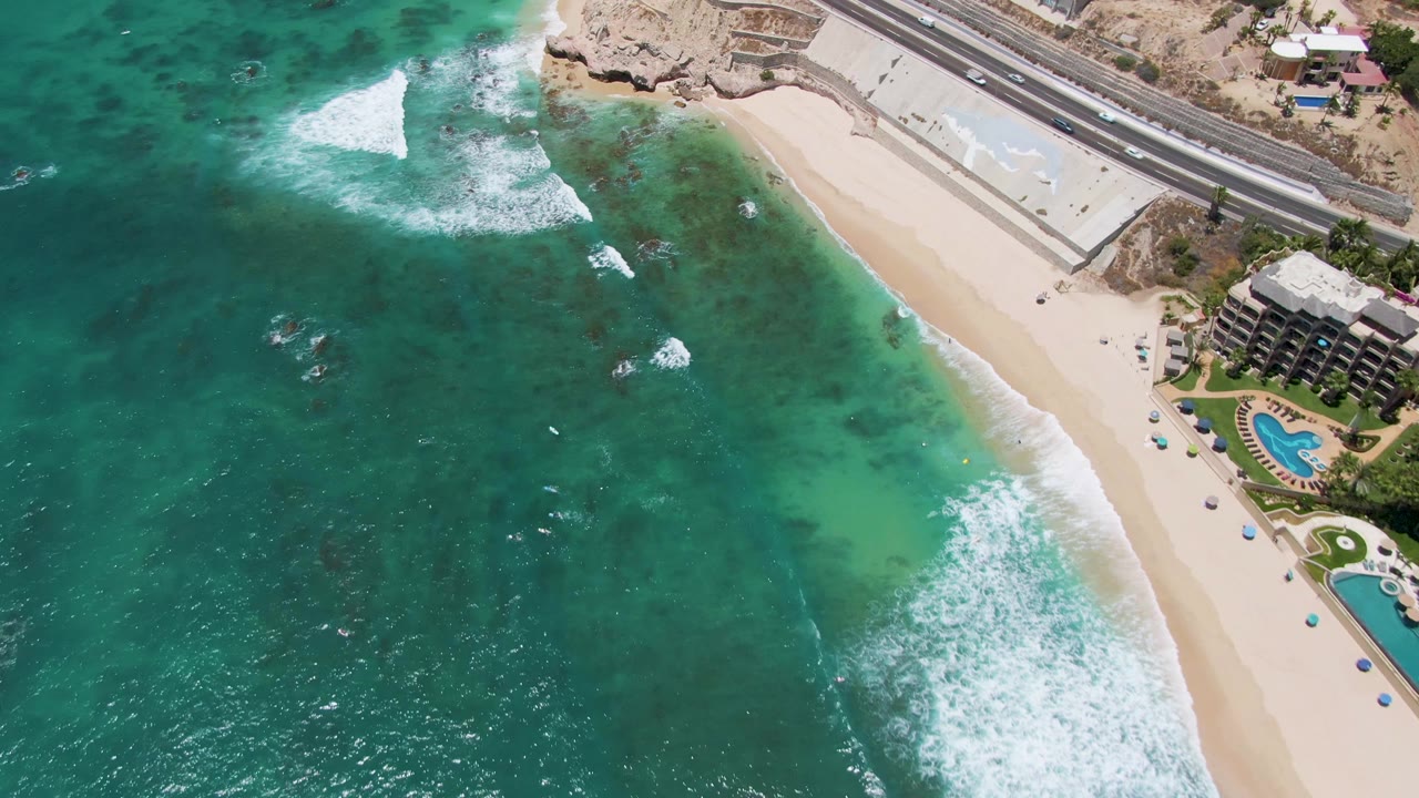 Drone view of clear green-blue water beside a sandy shoreline and road.