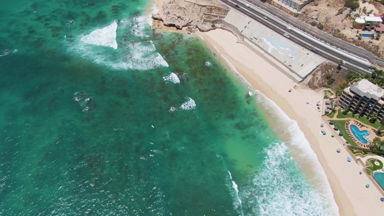 Aerial frame over Costa Azul Beach in Los Cabos where white water spreads near offshore rocks while the sandy beach, coastal road, buildings, pools, and no visible people remain in view.