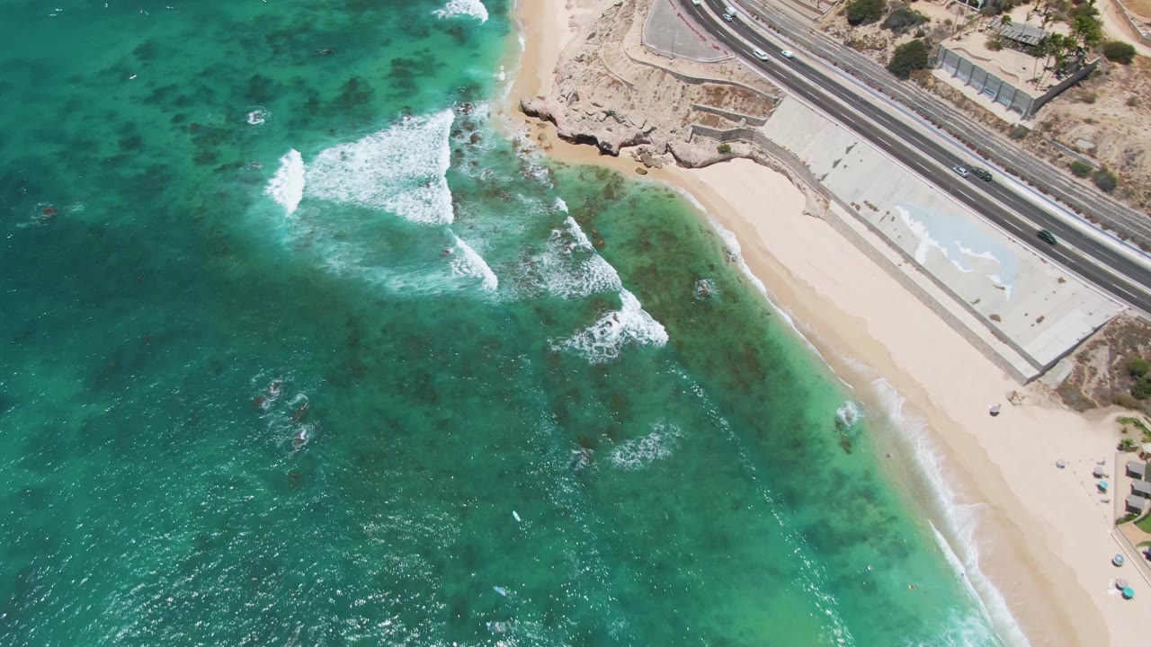 A high overhead view of Costa Azul Beach shows turquoise water, white surf near the sand, a parallel coastal road, and no visible people.