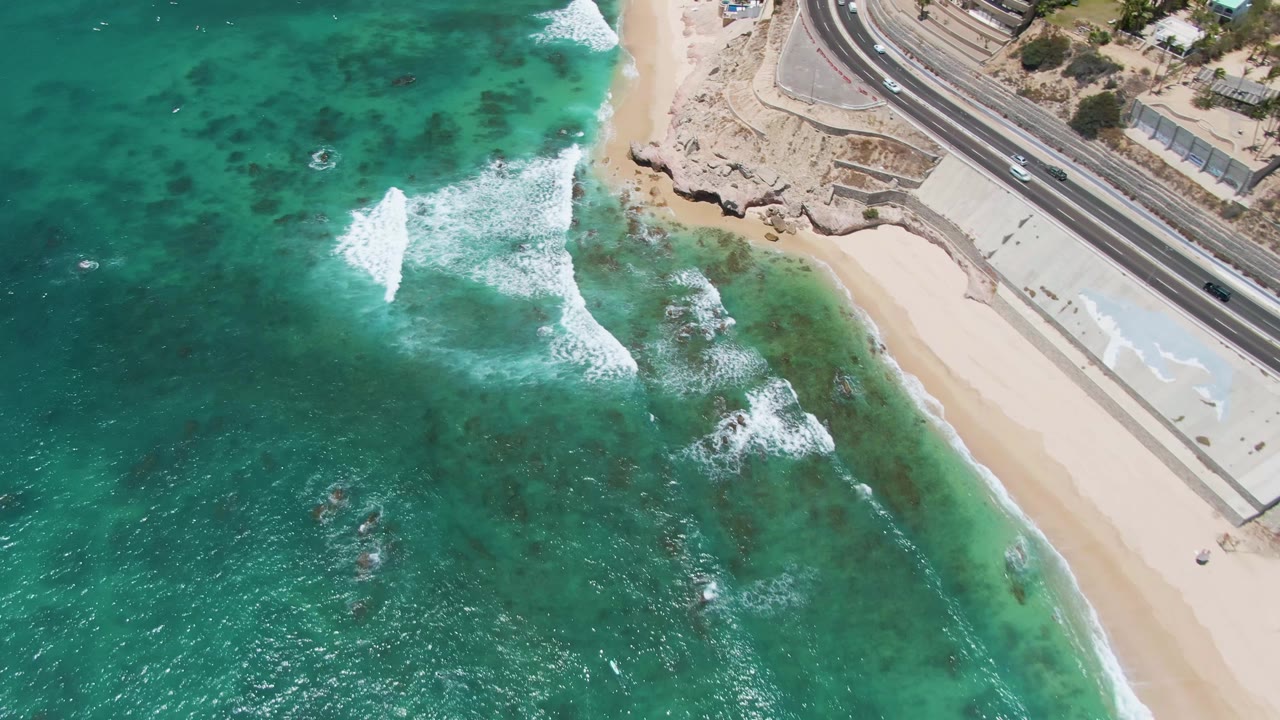 At 0:02, the wave pattern sits farther across the reef-colored water, with Costa Azul Beach on the right and the coastal road carrying vehicles in the upper frame, with no visible people.
