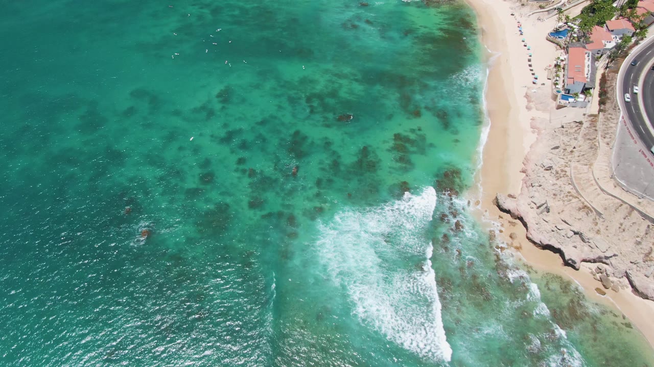 Overhead drone frame at Costa Azul Beach in Los Cabos showing turquoise water, dark seabed patches, white surf near the sandy shore, coastal buildings at the top right, and no discernible people.