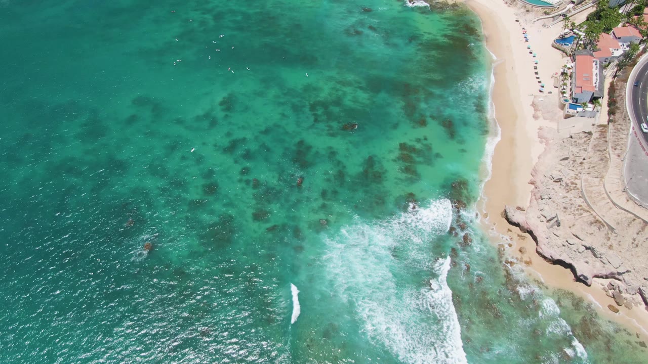 Costa Azul Beach aerial frame with clear turquoise shallows across the center, white foam pushing toward the lower right shore, rocks under the water, and no discernible people.