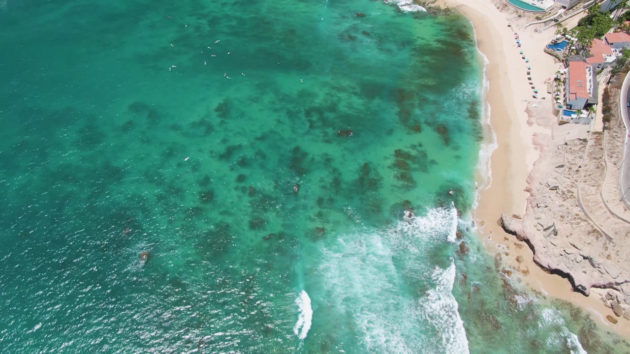 Overhead view at Costa Azul Beach with a broader patch of white surf crossing the lower right shallows, pale sand along the edge, and coastal structures clipped at the upper right with no discernible people.
