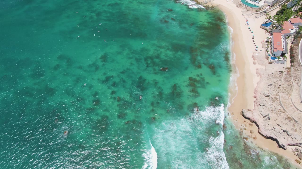 Top-down coastline view with green-blue water, foam, sand, and rocks.