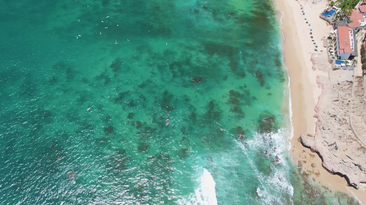 Overhead view at Costa Azul Beach in Los Cabos showing turquoise water, underwater rocks, a sandy shoreline, small coastal structures at the top edge, and no visible people.