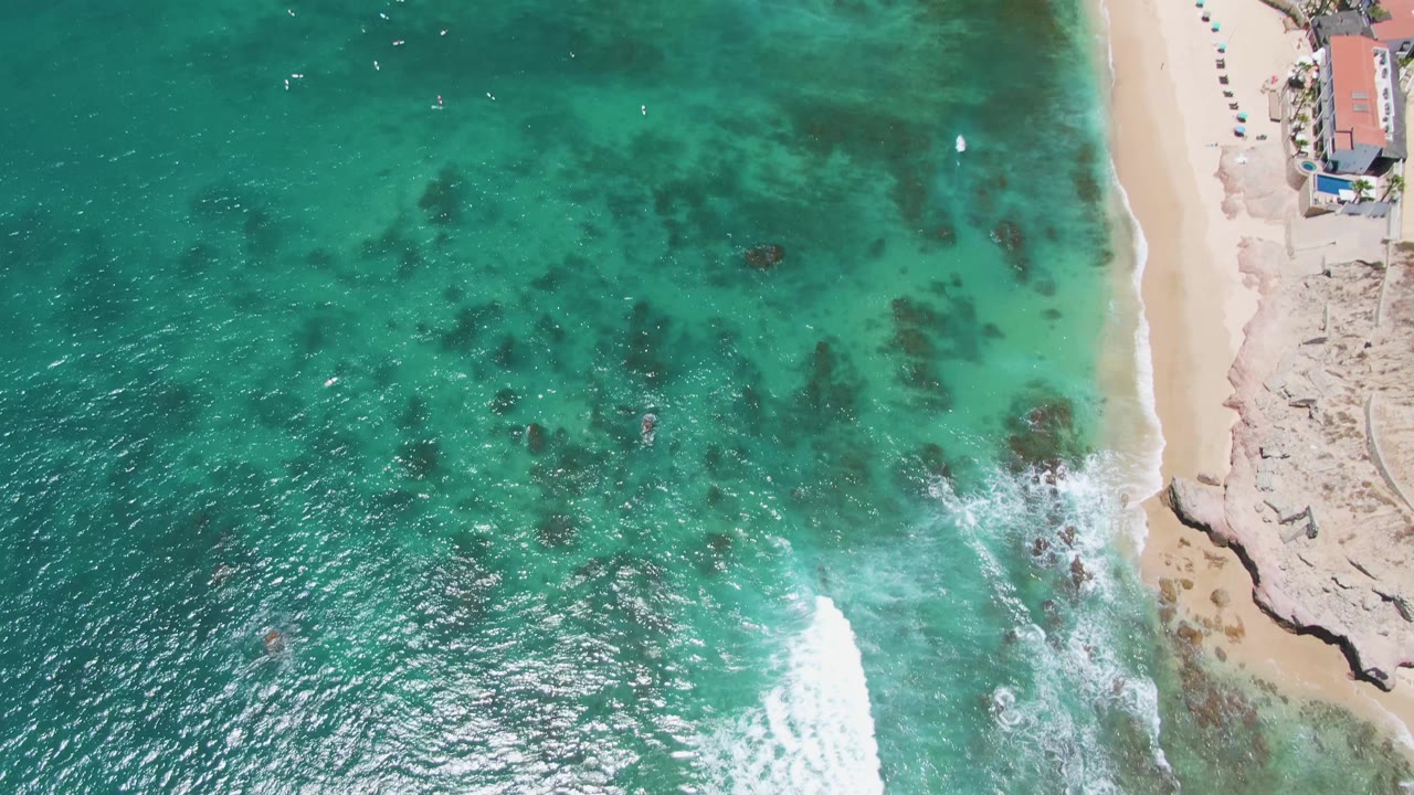 Overhead Costa Azul Beach frame with the wave line shifting closer to the sand, darker rocks visible under clear green-blue water, and no visible people.