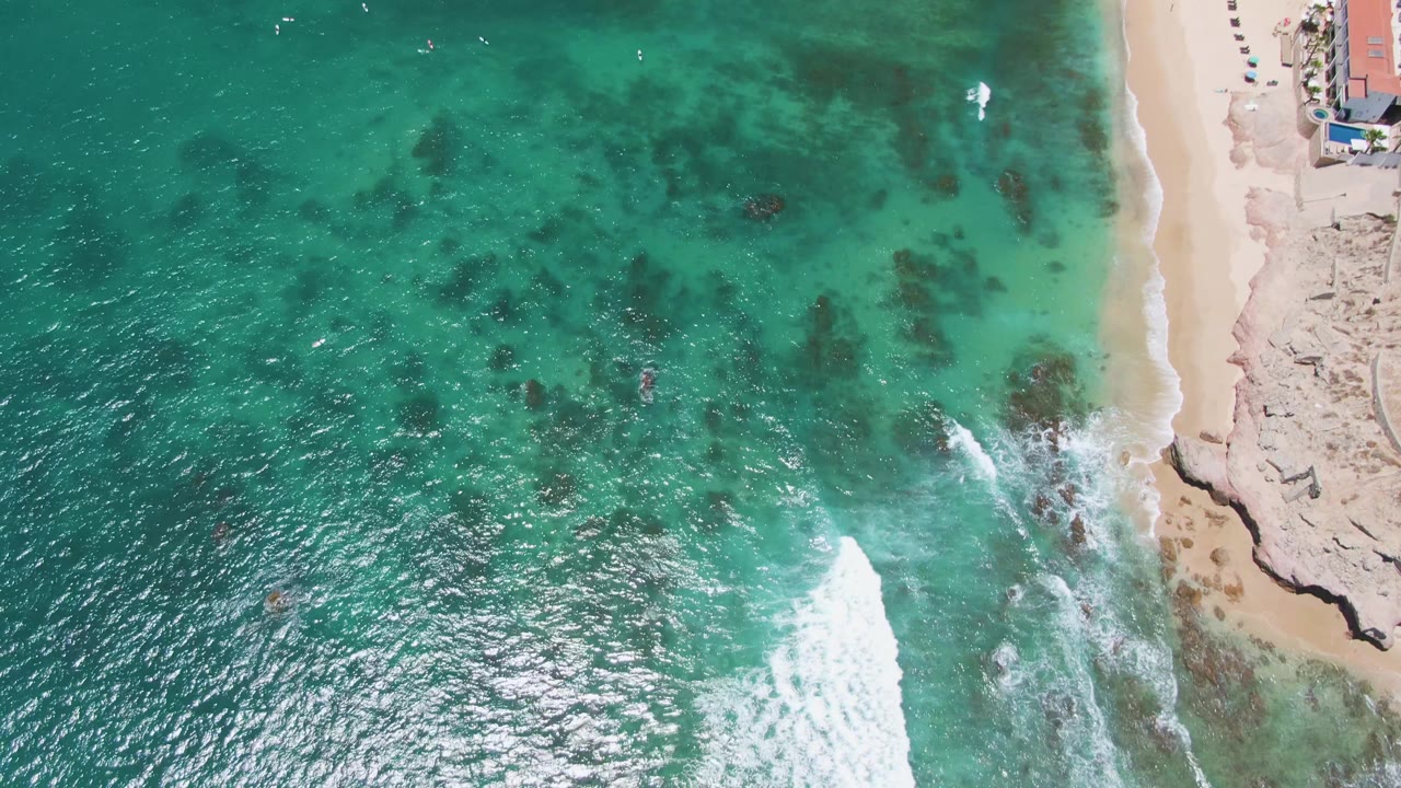 Aerial frame over Costa Azul Beach with bright foam spreading along the right-side rocks, sandy beach above it, clear turquoise water across the frame, and no visible people.