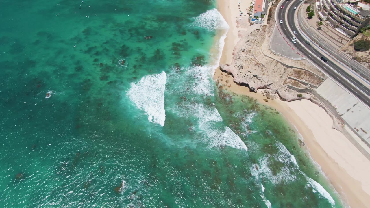 Overhead frame at Costa Azul Beach in Los Cabos shows turquoise water, white surf, sandy beach, rocky shoreline, and a curved road with cars, with no visible people.