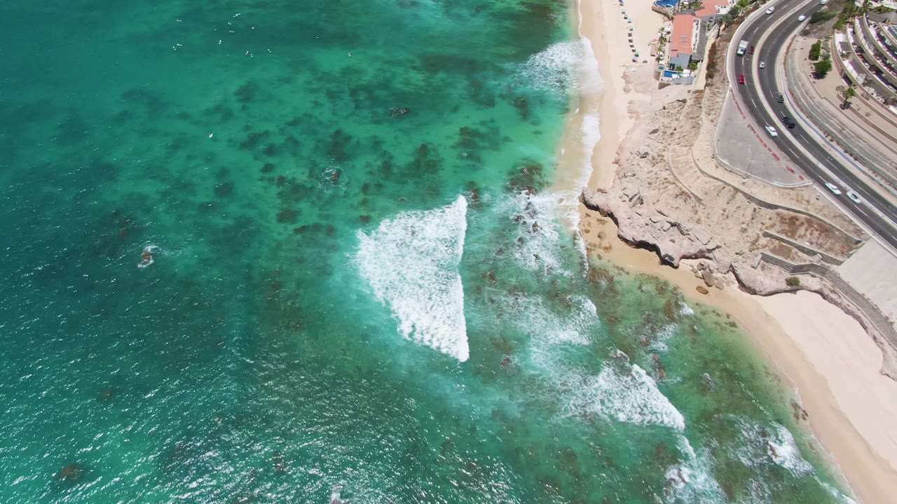 At 0:02, the drone view over Costa Azul Beach shows foamy surf closer to the rocky right shoreline, clear turquoise water, sand, and cars on the curved coastal road, with no visible people.