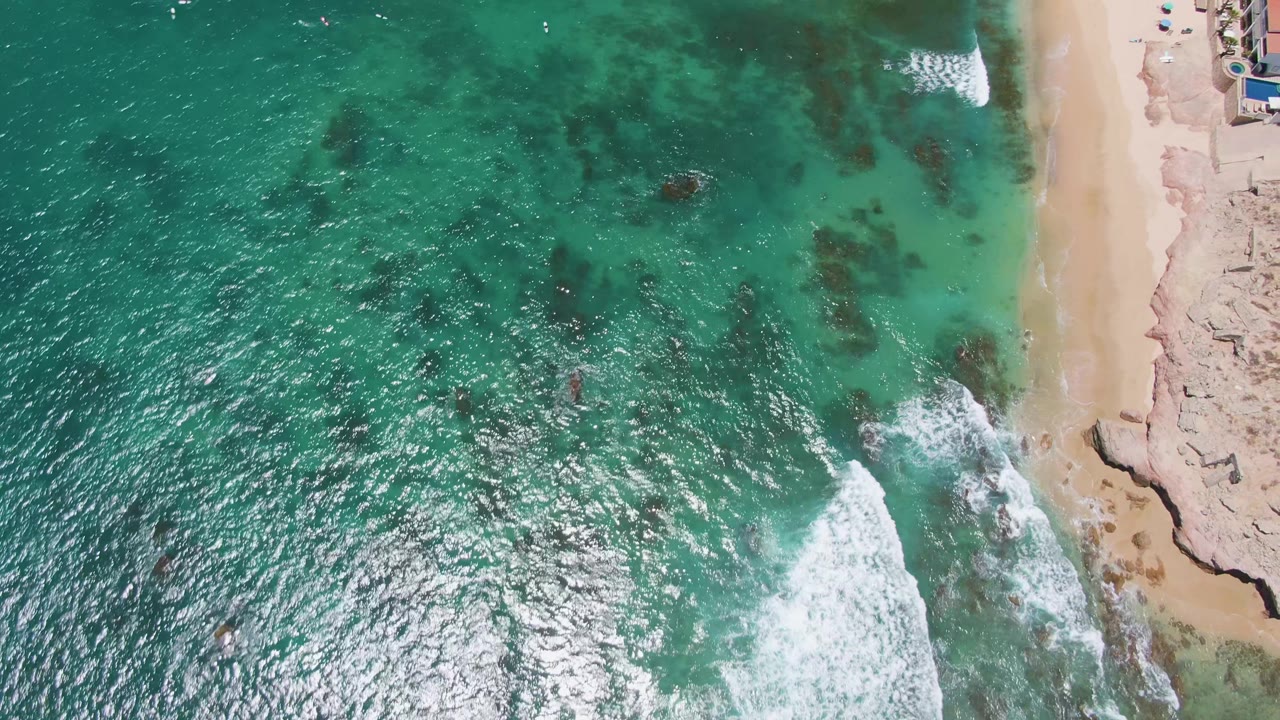Overhead view at Costa Azul Beach in Los Cabos showing turquoise water, dark submerged patches, white surf, sandy shore, rocky outcrops, and no visible people.