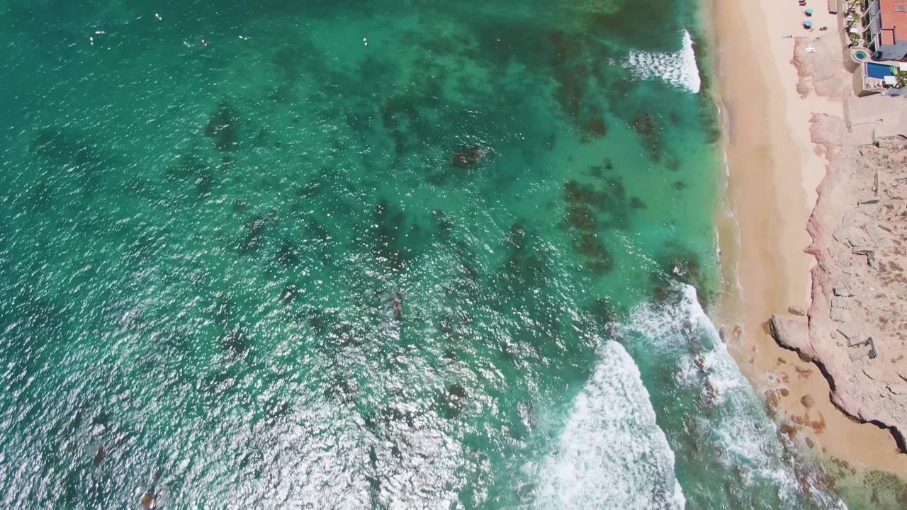 Aerial view of waves spreading across clear water toward a narrow beach.