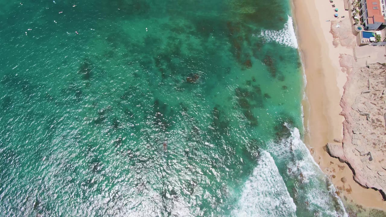 At 0:02, sunlight sparkles across turquoise water at Costa Azul Beach, with dark underwater patches offshore and surf curling near the sandy beach, no visible people.