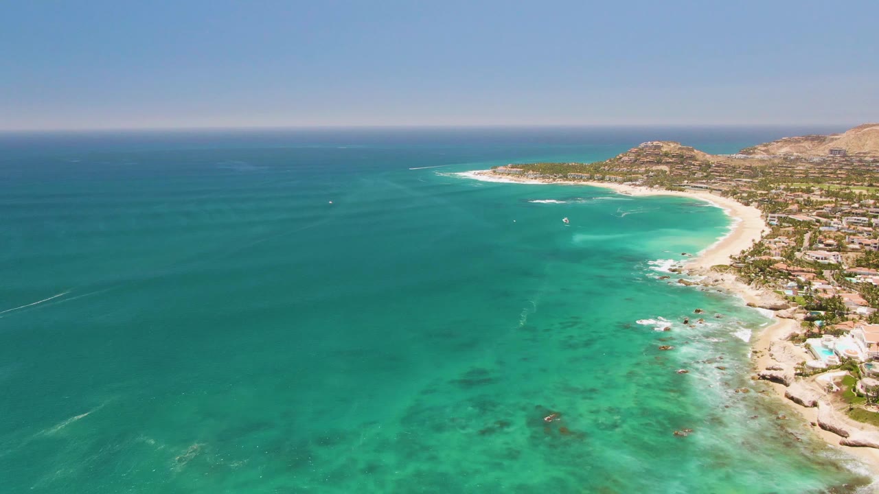 Aerial frame over Costa Azul Beach in Los Cabos with turquoise water across the foreground, a curved sandy shoreline on the right, coastal buildings beyond the beach, and no visible people.