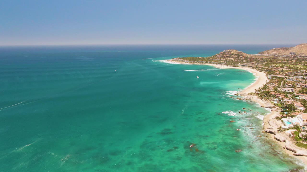 High aerial view at Costa Azul Beach, with blue-green ocean filling the left side, pale surf tracing the beach on the right, clustered coastal buildings inland, and no visible people.