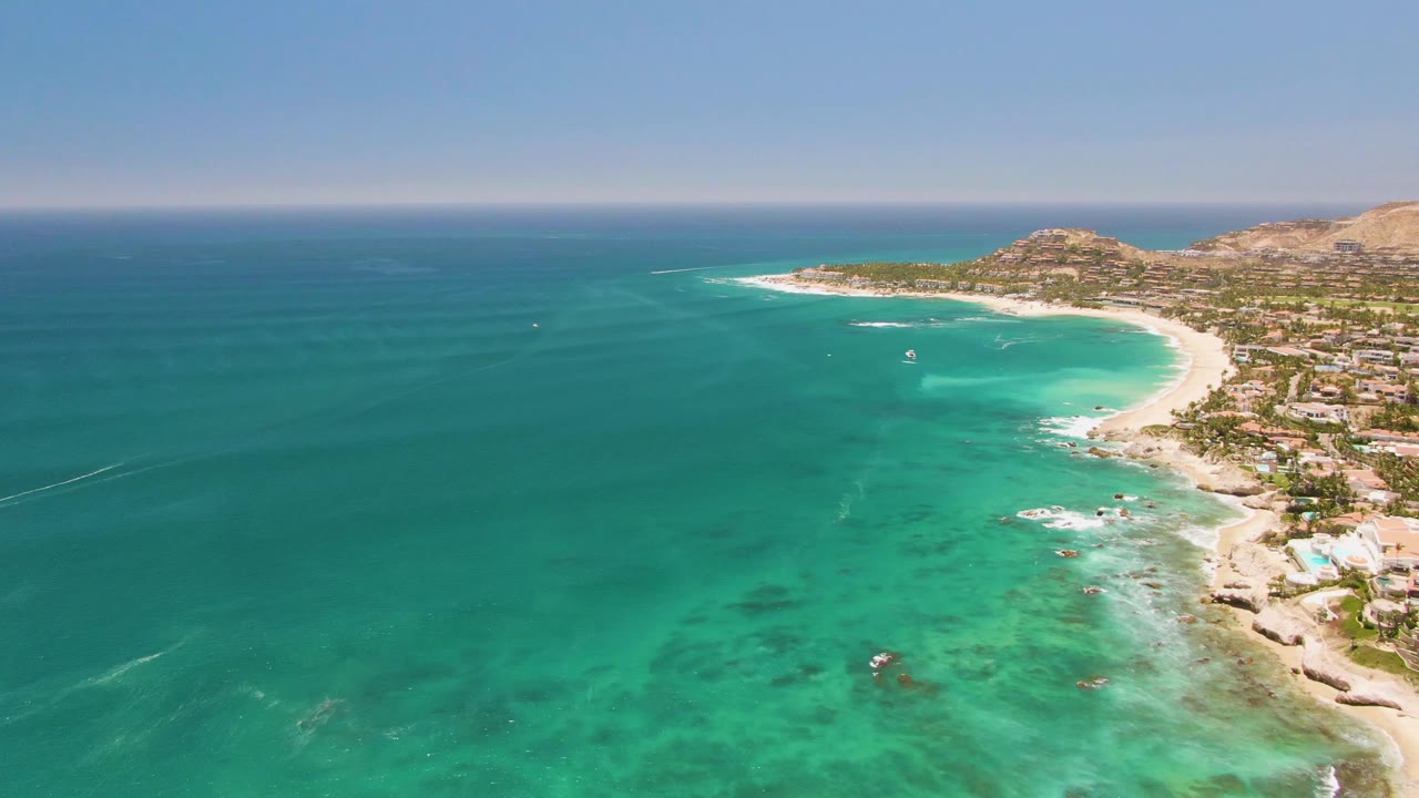 Wide aerial coastline with turquoise water, sand, and low buildings.
