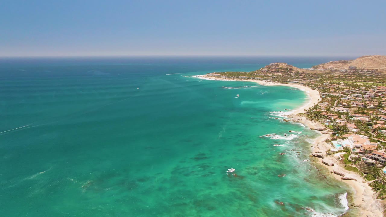 Aerial view over Costa Azul Beach in Los Cabos with turquoise water, a curved sandy shoreline, beachfront buildings, distant hills, and no visible people.