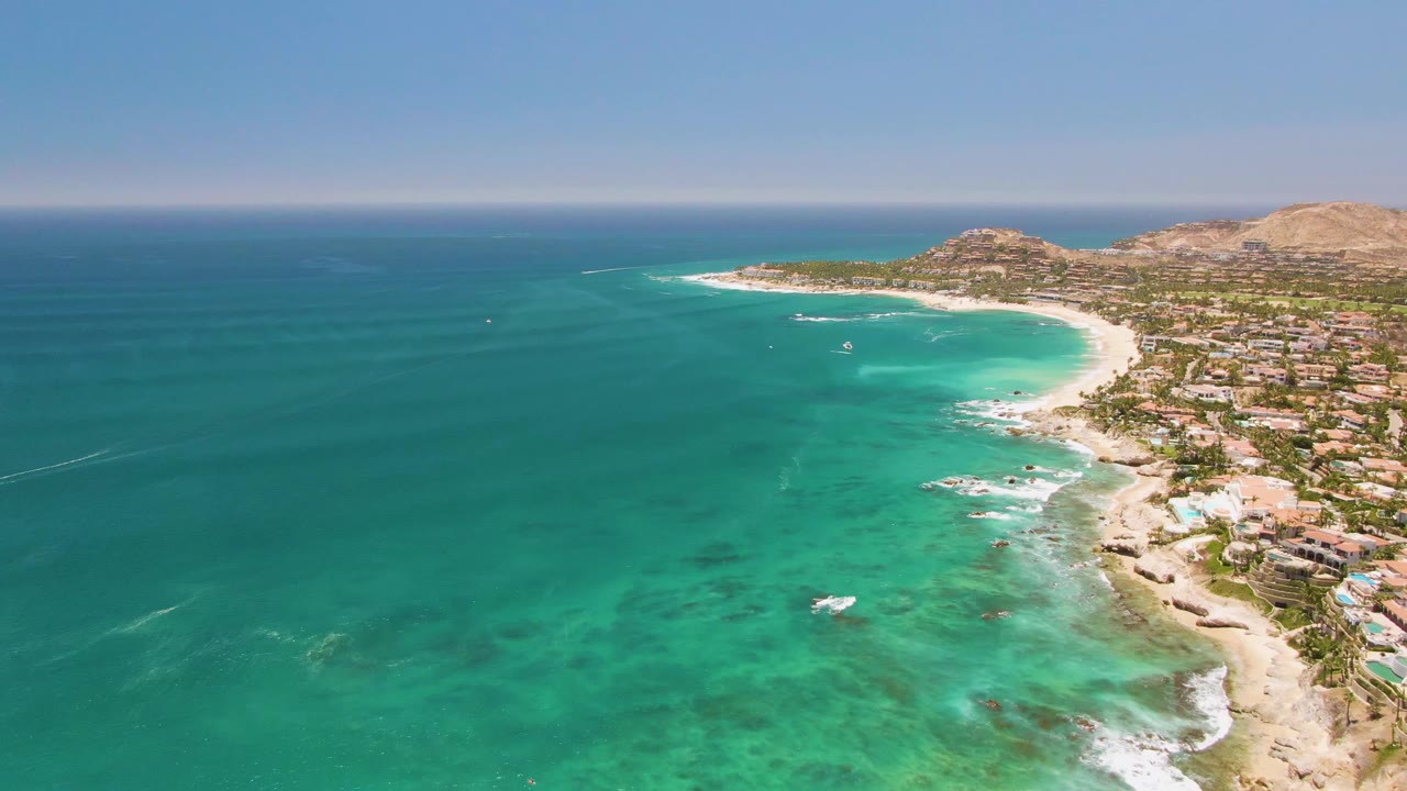 Aerial view of Costa Azul Beach with bright turquoise shallows near the sand, small white breakers, coastal buildings at right, dry hillside terrain beyond, and no visible people.