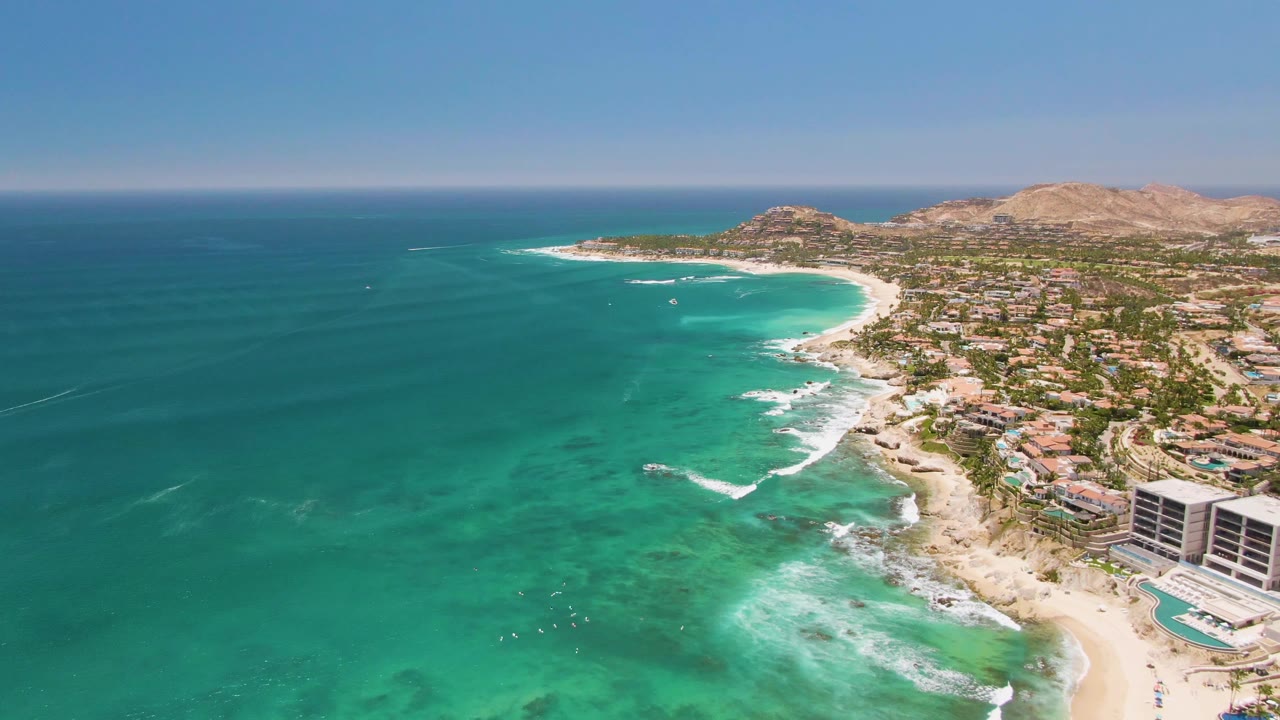 Aerial frame over Costa Azul Beach in Los Cabos showing turquoise water on the left, white surf along the sand, coastal buildings on the right, and low hills beyond the shoreline.