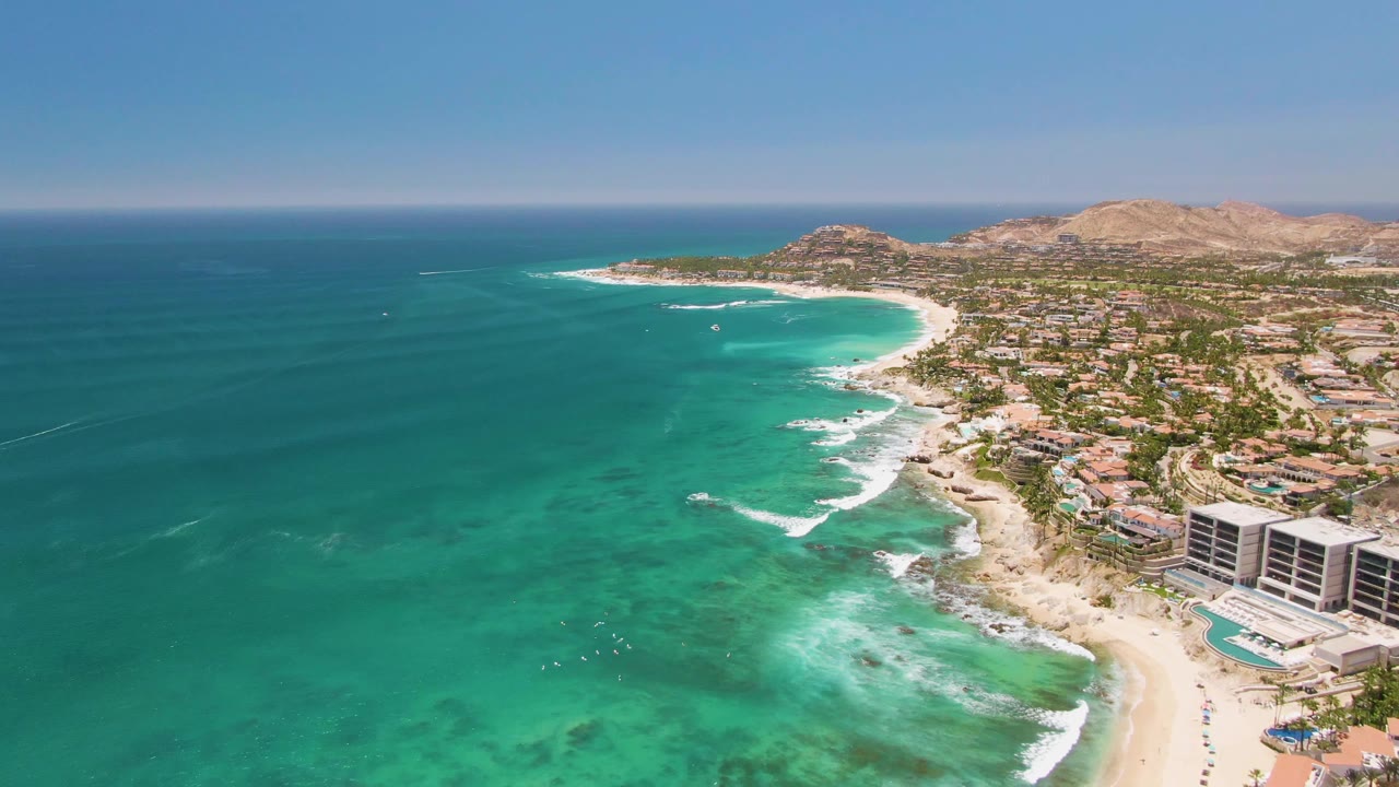 Wide drone frame over Costa Azul Beach showing clear green-blue water with darker reef patches, surf bands meeting the sand, beachfront buildings at right, and dry hills behind town.