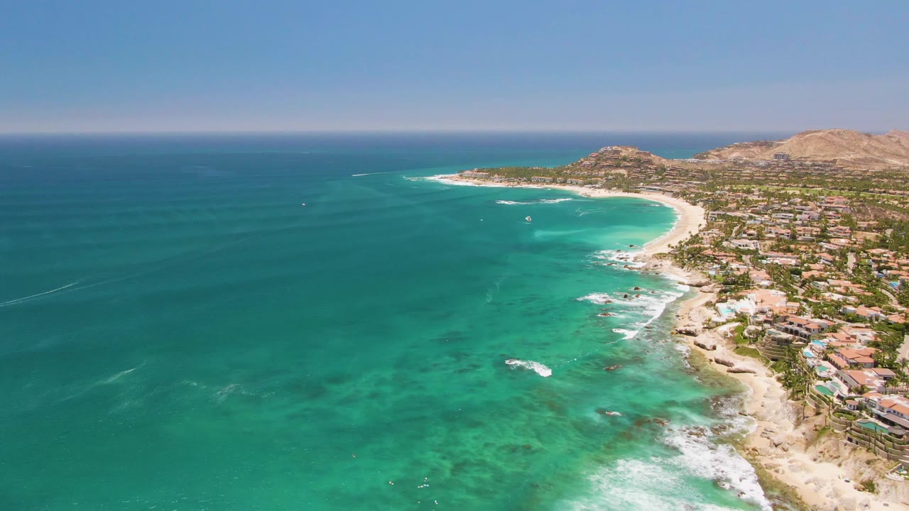 Opening aerial frame over Costa Azul Beach in Los Cabos, with turquoise water at left, white surf on the sand, coastal buildings to the right, dry hills beyond, and no visible people.