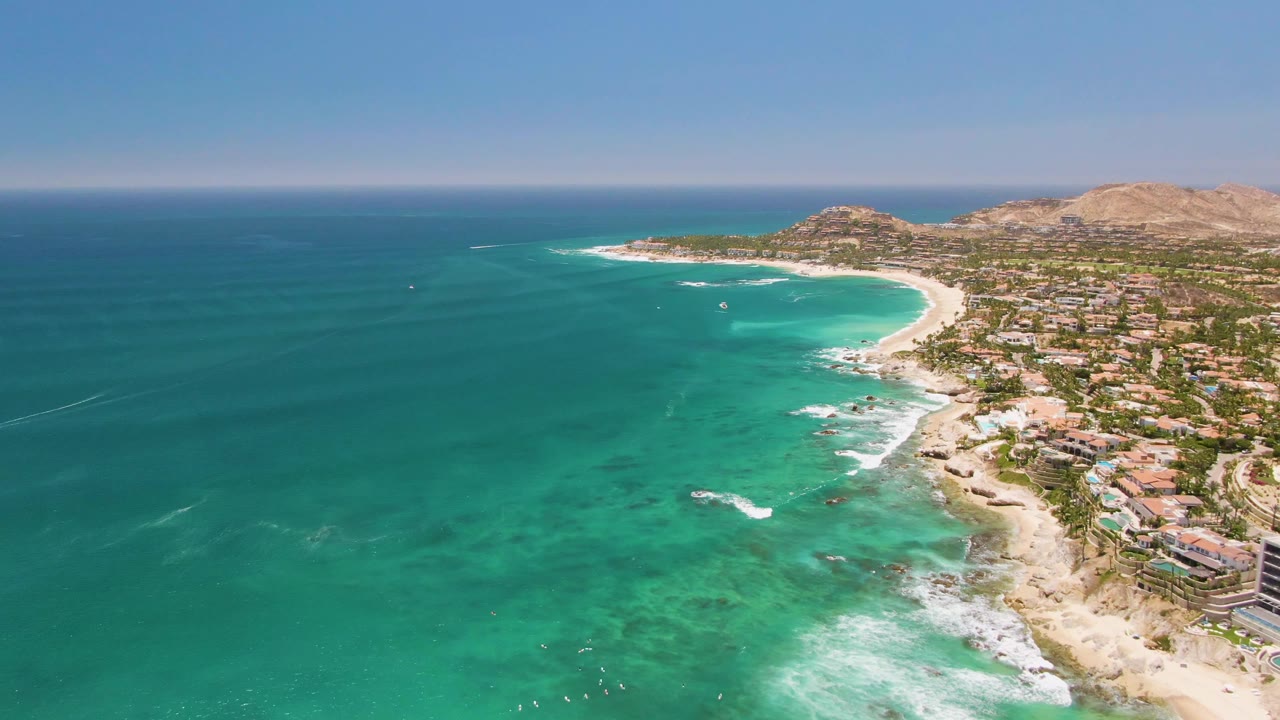 Mid-clip aerial view over Costa Azul Beach, with darker ocean water offshore, pale turquoise shallows, surf curling toward sand, buildings lining the coast, hills in the background, and no visible people.