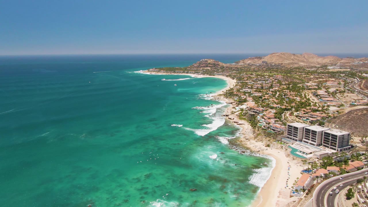 Opening aerial frame over Costa Azul Beach in Los Cabos, with turquoise water at left, surf along the sand, coastal buildings and a curved road at right, and no visible people.