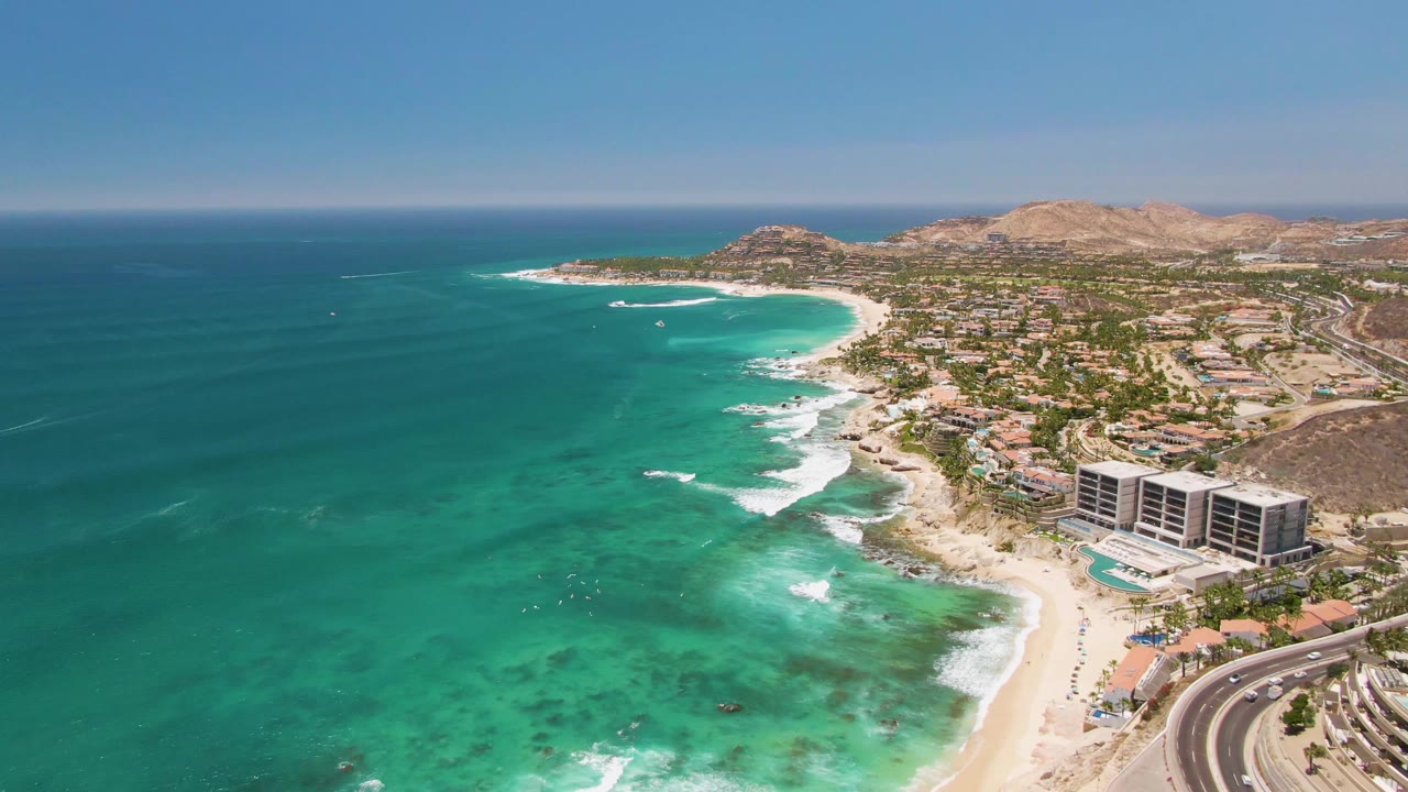 Aerial frame over Costa Azul Beach in Los Cabos showing the shoreline slightly farther along the curve, with white waves near the sand, traffic on the road, buildings, hills, and no visible people.