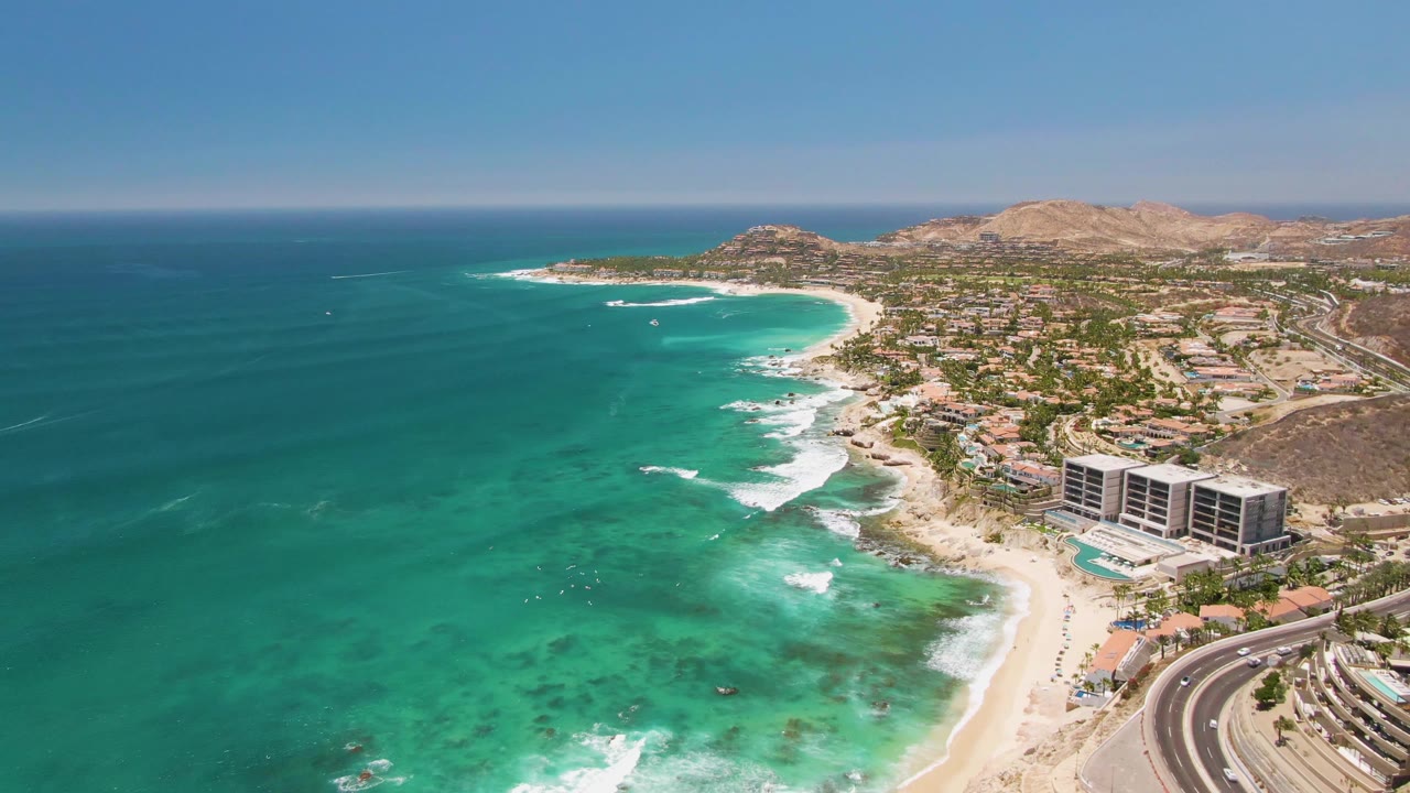 Turquoise bay with surf, beach, buildings, curved road, and arid hills beyond.