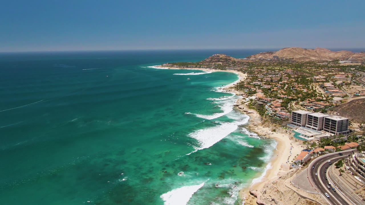 Aerial frame over Costa Azul Beach in Los Cabos showing turquoise water at left, white surf on the sand, a curved coastal road at lower right, beachfront buildings, dry hills, and no visible people.