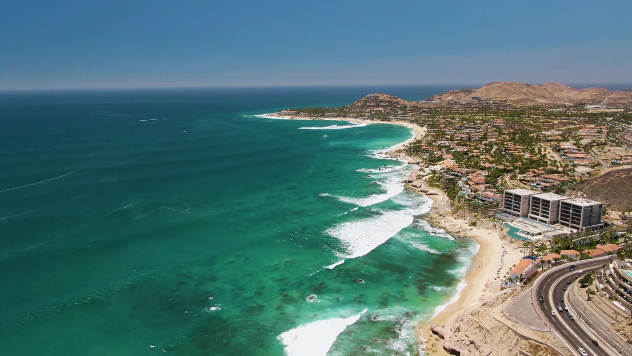 Wide drone view of turquoise water and white surf beside a beach road with vehicles.