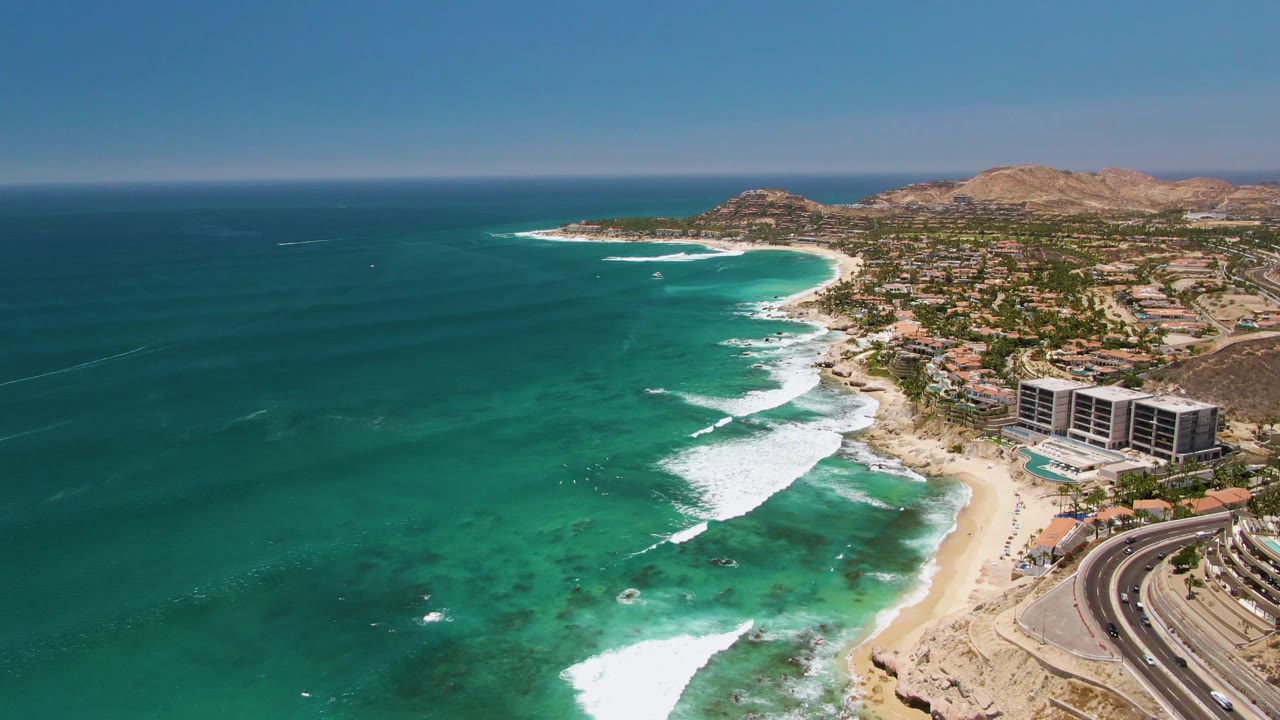 Costa Azul Beach aerial frame showing teal ocean water across the left half, staggered white breakers near shore, a sandy cove, a roadside curve with vehicles, nearby buildings, and no visible people.