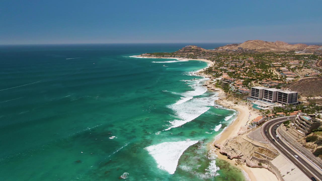 Wide aerial frame of Costa Azul Beach in Los Cabos with turquoise water, white surf, sandy shore, coastal buildings, a highway, dry hills, and no visible people.