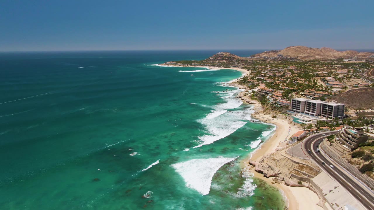 Turquoise surf rolls toward a sandy beach near coastal buildings.